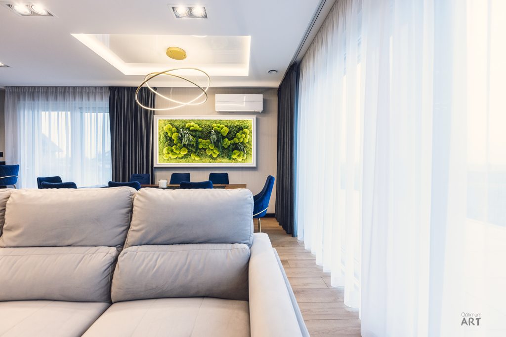 Nowoczesny salon z jasną sofą na pierwszym planie i jadalnią z niebieskimi krzesłami w tle. W centrum uwagi znajduje się duży obraz z zieloną roślinnością na ścianie oraz złota lampa wisząca nad stołem jadalnym.