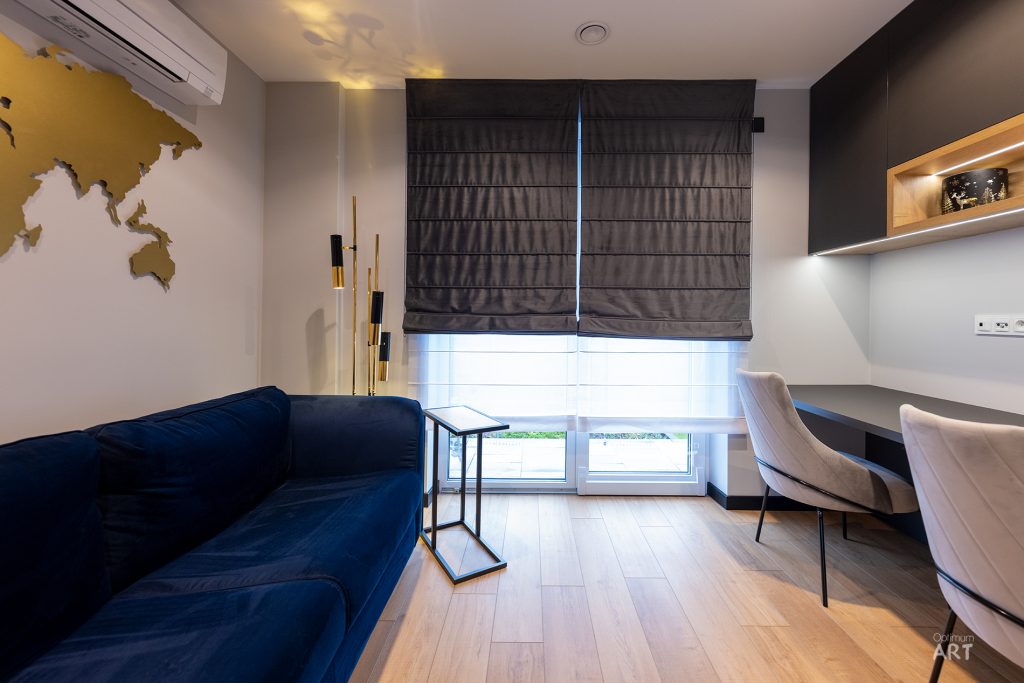 Nowoczesne mieszkanie z granatową sofą, białymi krzesłami przy biurku oraz dużym oknem z szarymi roletami. Na ścianie znajduje się złota mapa świata, a pomieszczenie ma jasne, drewniane podłogi.