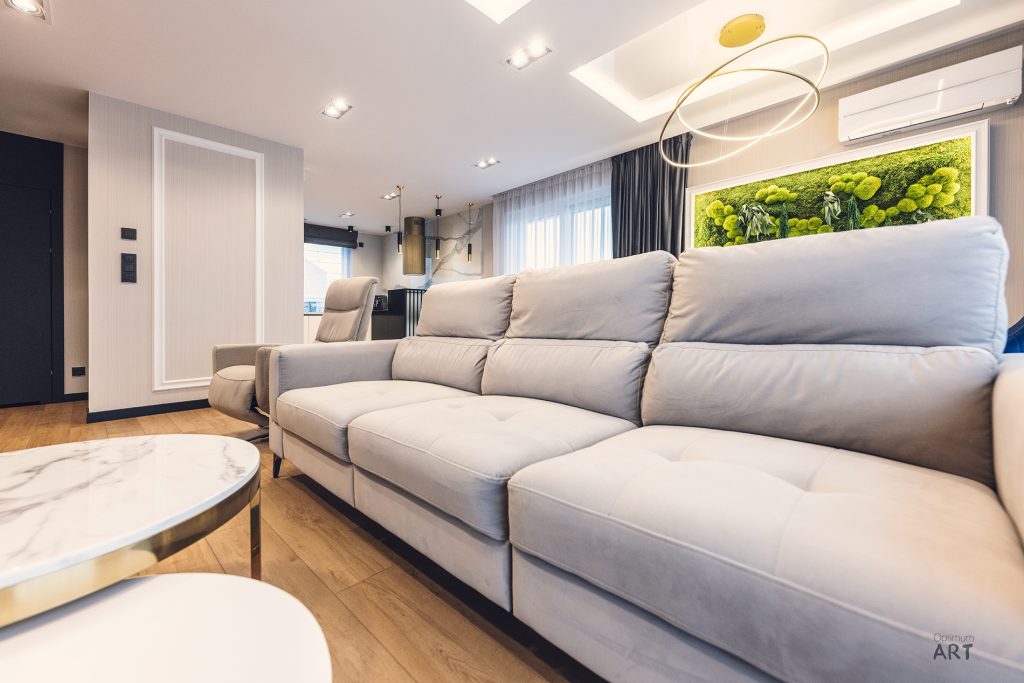 Nowoczesny salon z dużą szarą sofą, okrągłym stolikiem kawowym i dekoracyjną lampą wisząca. W tle widoczna kuchnia z wyspą oraz zielona dekoracja roślinna na ścianie.