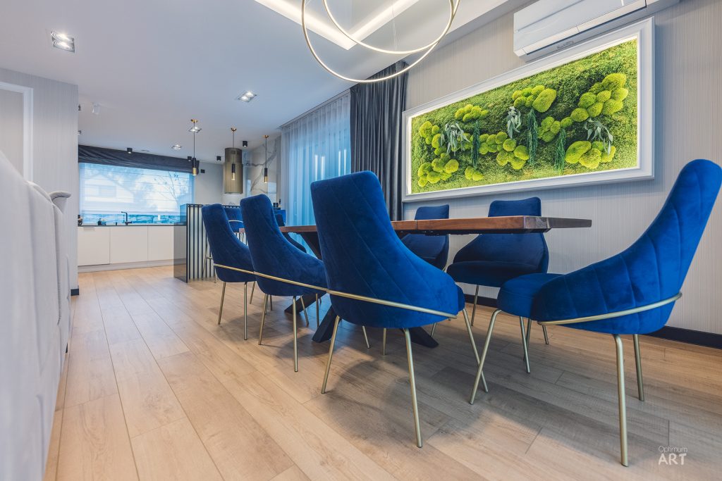 Nowoczesna jadalnia z okrągłym drewnianym stołem otoczonym sześcioma niebieskimi krzesłami o welurowym wykończeniu. Na ścianie wisi duży obraz z zielonym motywem roślinnym, a w tle widać otwartą kuchnię z białymi meblami.