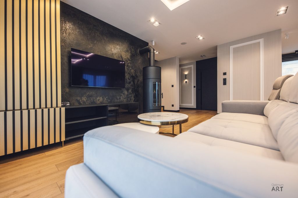 Nowoczesny salon z jasną kanapą, drewnianą podłogą i telewizorem zamontowanym na ciemnej ścianie z teksturą. Po lewej stronie widoczna jest drewniana ścianka z pionowymi listwami, a w centrum pomieszczenia stoi okrągły stolik kawowy.