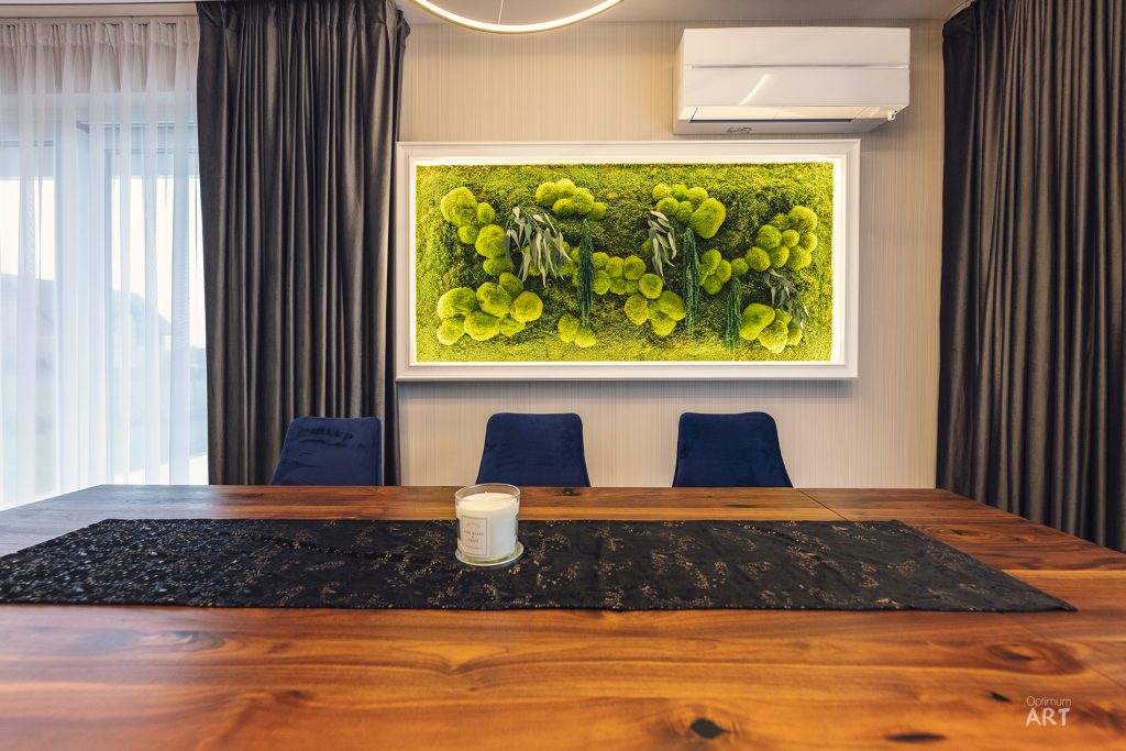 Nowoczesna jadalnia z drewnianym stołem, na którym stoi świeca, oraz trzema granatowymi krzesłami. Na ścianie wisi duży obraz z zielonymi mchem i roślinami, a okna zasłaniają ciemne zasłony.