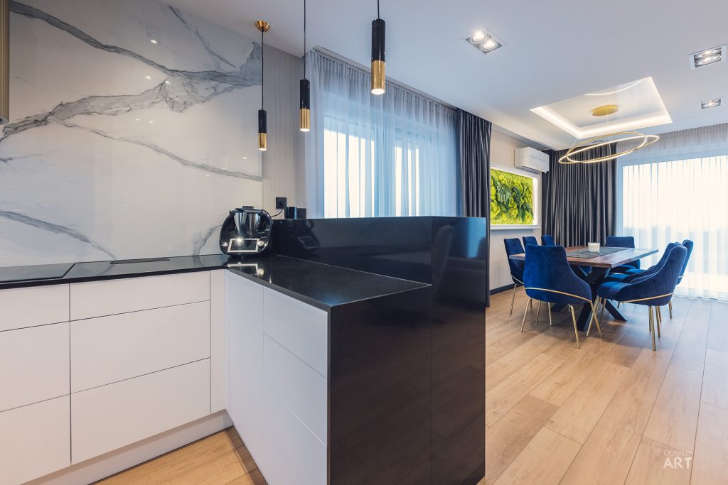 Nowoczesna kuchnia z białymi szafkami dolnymi, czarnym blatem i marmurowymi ścianami, połączona z jadalnią z niebieskimi krzesłami wokół okrągłego stołu. Wnętrze ma eleganckie oświetlenie wiszące i duże okna z zasłonami.