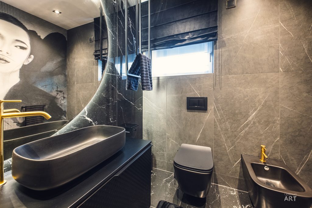Nowoczesna łazienka z ciemnymi marmurowymi płytkami, owalna umywalka na czarnej szafce, toaleta i bidet. Na ścianie wisi duży portret kobiety, a przez okno z żaluzjami wpada naturalne światło.