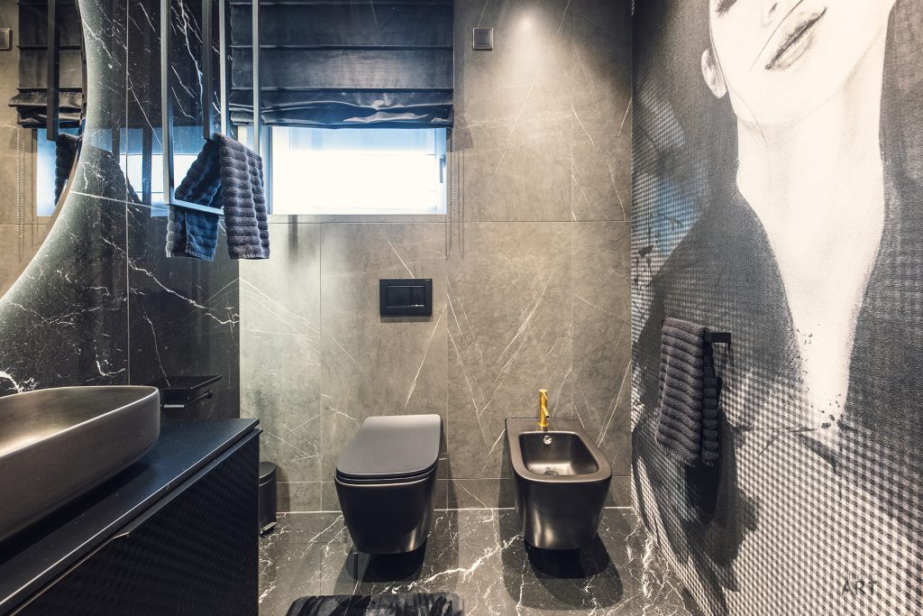Nowoczesna łazienka z ciemnymi marmurowymi płytkami, w której znajdują się toaleta, bidet, umywalka na blacie oraz kabina prysznicowa. Na ścianie widoczna jest duża czarno-biała grafika przedstawiająca twarz kobiety.