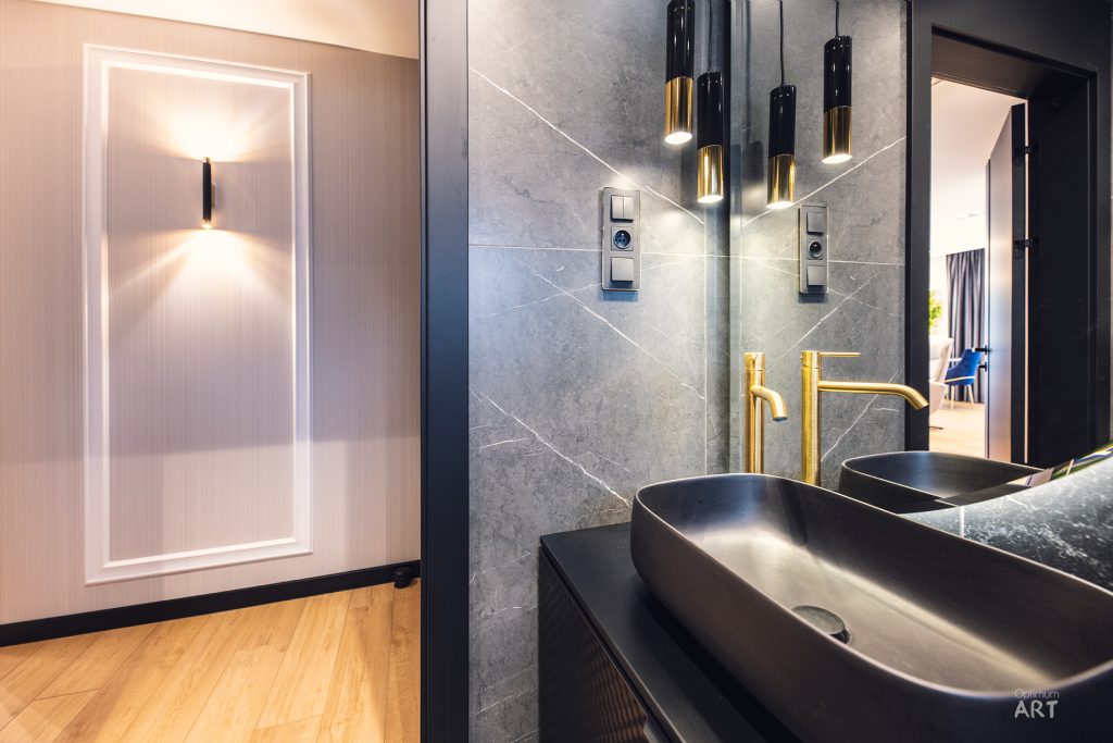 Nowoczesna łazienka z dwoma czarnymi umywalkami nablatowymi, złotymi bateriami i wiszącymi lampami. Na ścianie znajduje się duże lustro, a po lewej stronie widoczne jest podświetlone lustro na jasnej ścianie z drewnianą podłogą.