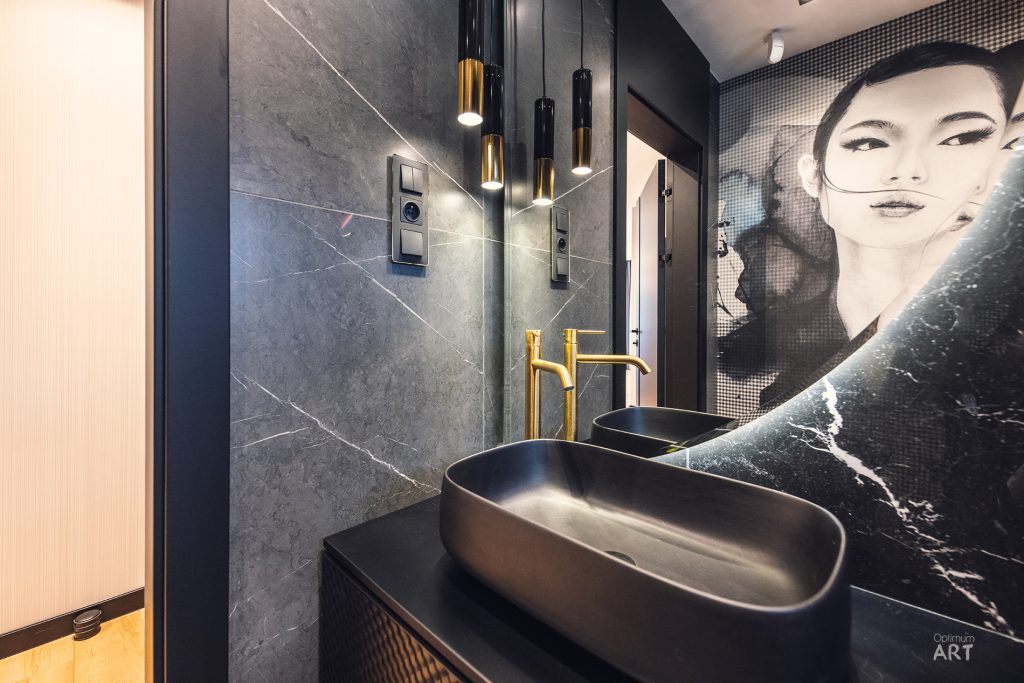Nowoczesna łazienka z czarnymi owalnymi umywalkami na blacie, złotymi bateriami i lampami wiszącymi. Na ścianie widoczny jest artystyczny portret kobiety w odcieniach szarości, a wnętrze utrzymane jest w eleganckich ciemnych kolorach z marmurową ścianą.