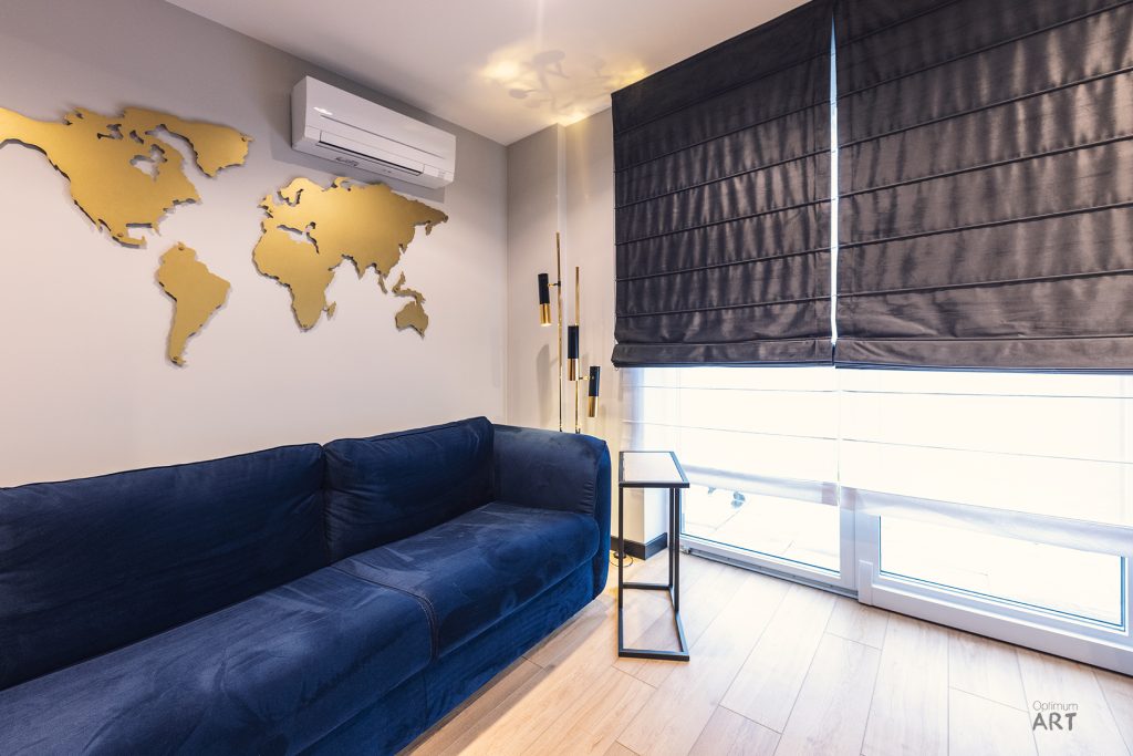 Nowoczesny salon z granatową narożną sofą, złotą mapą świata na białej ścianie i dużym oknem z szarymi roletami. Na podłodze stoi elegancka lampa podłogowa z geometryczną podstawą.