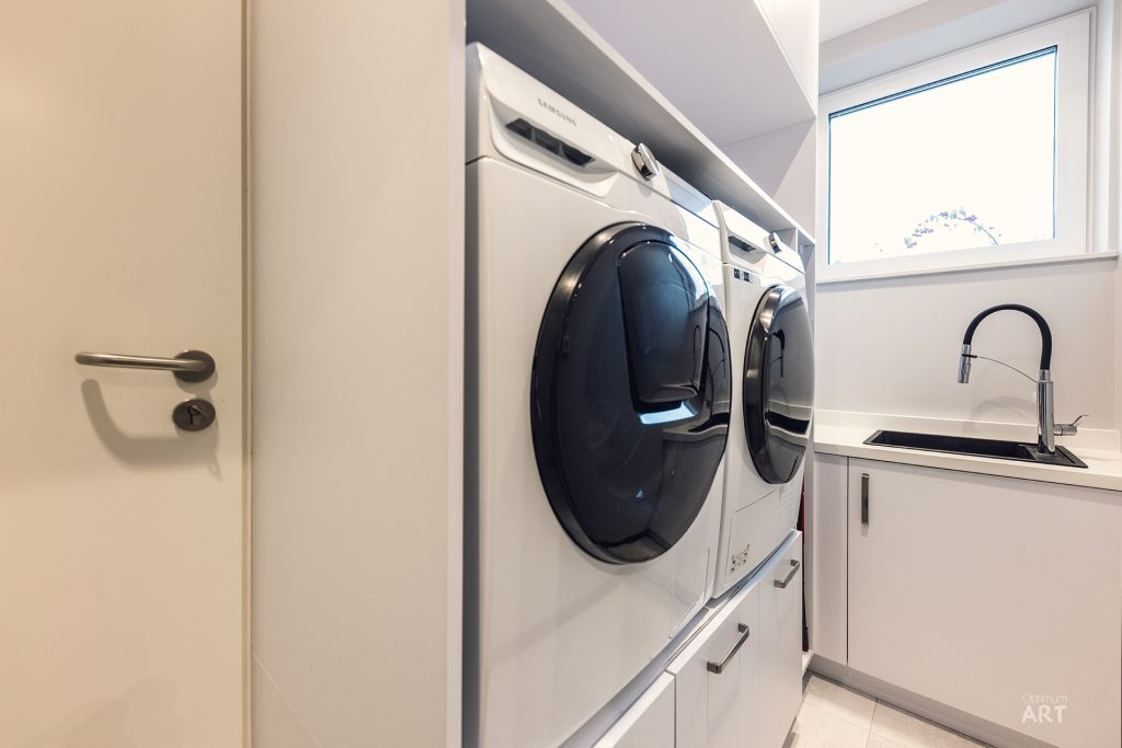 Nowoczesna pralnia z białymi meblami, w której znajdują się dwie pralki z okrągłymi, ciemnymi drzwiczkami oraz zlew z kranem. Po prawej stronie widać okno wpuszczające naturalne światło do pomieszczenia.