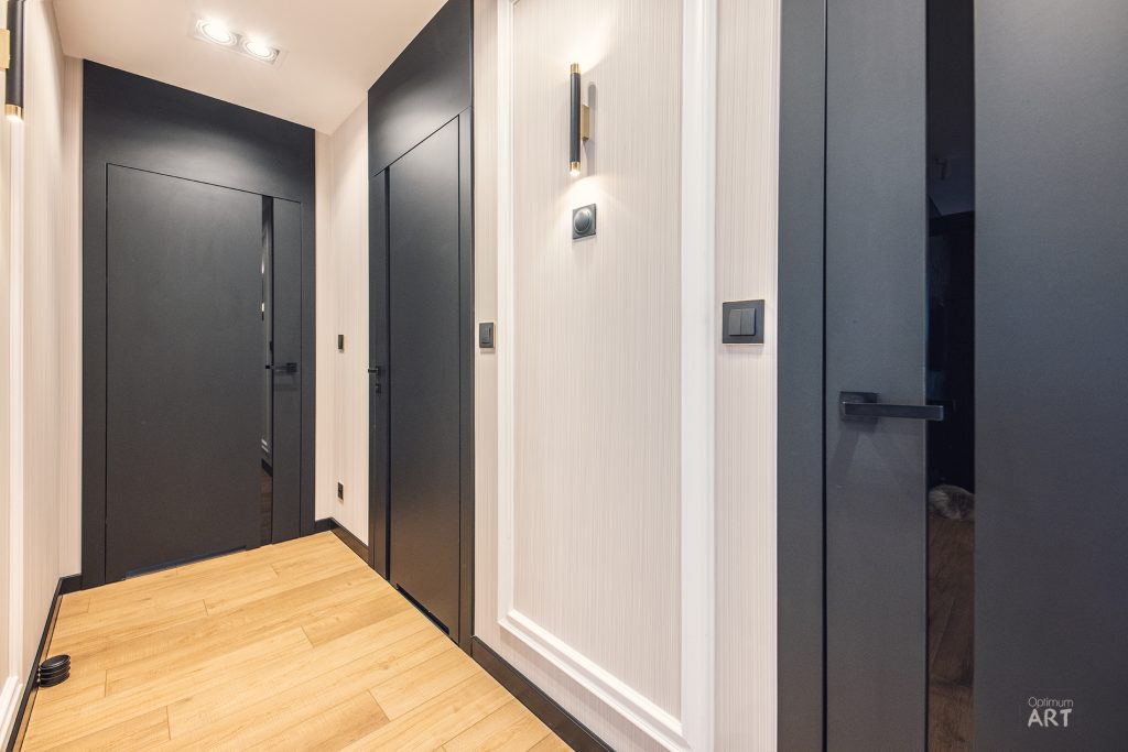 Nowoczesny korytarz mieszkania z jasnymi ścianami i ciemnymi drzwiami, z drewnianą podłogą i minimalistycznym oświetleniem. Po prawej stronie widoczne są otwarte drzwi prowadzące do ciemniejszego pomieszczenia.
