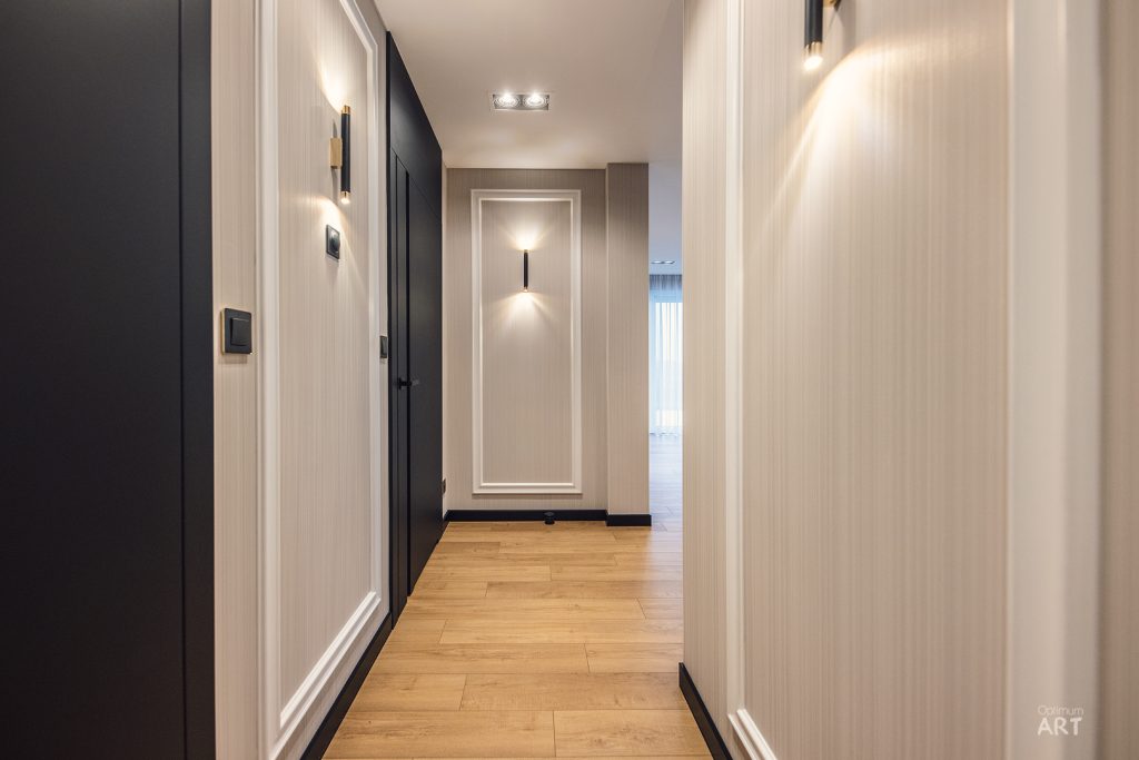Elegancki korytarz w nowoczesnym stylu z drewnianą podłogą, białymi ścianami po prawej stronie i czarnymi elementami po lewej. Przestrzeń jest dobrze oświetlona lampami ściennymi i sufitowymi, z widocznymi drzwiami na końcu korytarza.