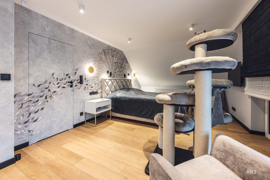 Nowoczesna sypialnia z łóżkiem o pikowanym zagłówku, szarą ścianą z dekoracyjnym wzorem i dużym drapkiem dla kota z beżowymi platformami. Pomieszczenie ma drewnianą podłogę, białe sufity z oświetleniem punktowym i minimalistyczne meble.