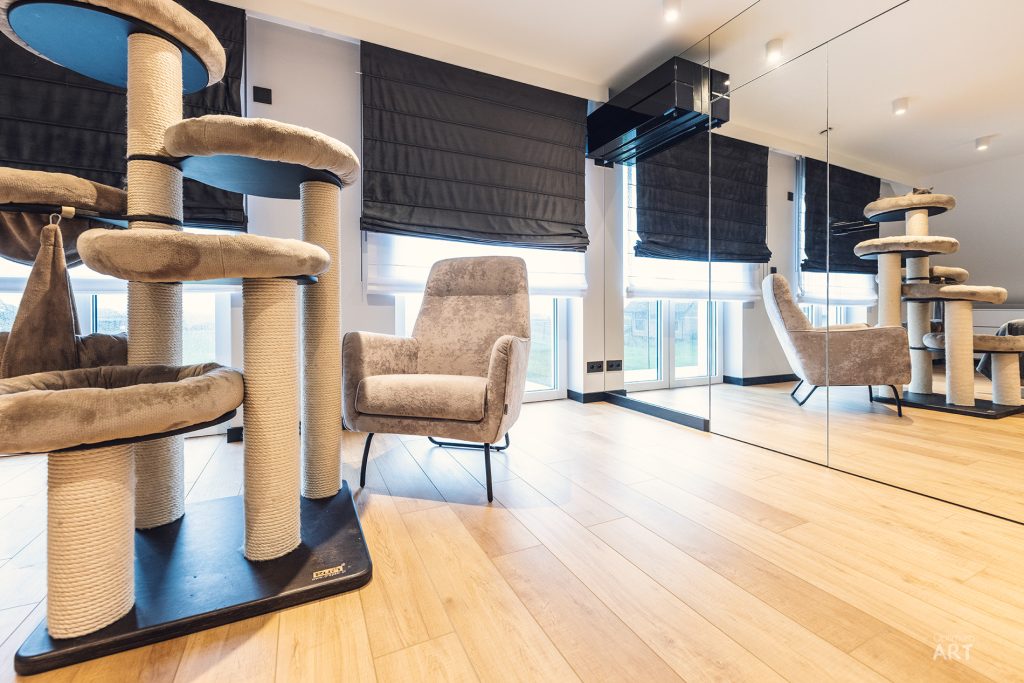 Nowoczesne wnętrze z dużymi drapakami dla kotów wykonanymi z sizalu, beżowym fotelem oraz lustrzaną ścianą. Pomieszczenie ma drewnianą podłogę, czarne rolety w oknach i białe ściany z nowoczesnym oświetleniem.