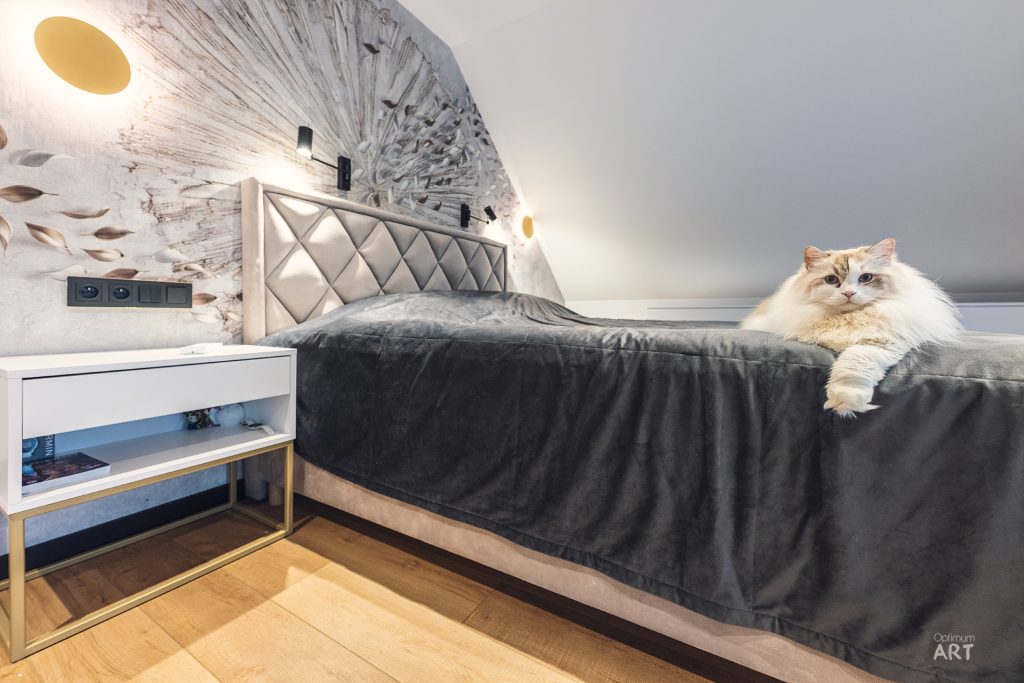 Nowoczesna sypialnia z łóżkiem z pikowanym zagłówkiem i szarą pościelą, na której siedzi puszysty kot o jasnym futrze. Wnętrze ma stylową dekorację ścienną z motywem dandeliona i drewnianą podłogę.