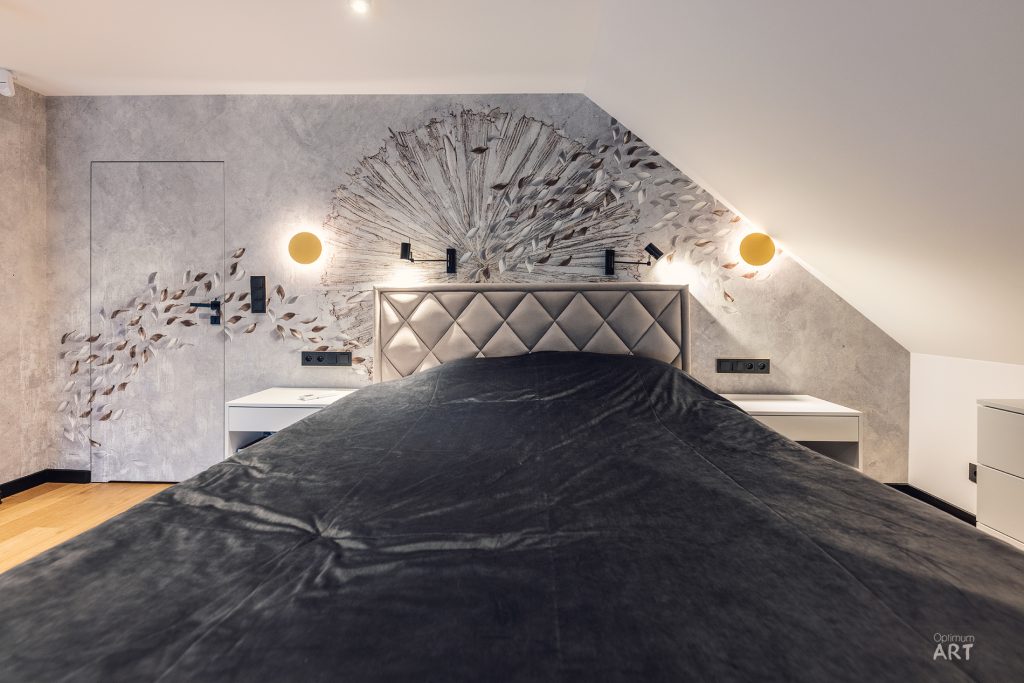 Nowoczesna sypialnia z łóżkiem z czarną pościelą i pikowanym wezgłowiem, umieszczona pod skośnym sufitem. Na ścianie za łóżkiem znajduje się dekoracyjna rzeźba przypominająca eksplodujące elementy, a po bokach świecą okrągłe lampki ścienne.
