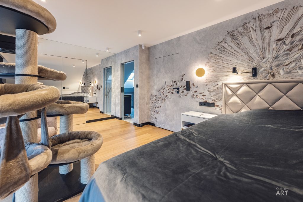 Nowoczesna sypialnia z dużym łóżkiem z szarą pościelą i tapicerowanym zagłówkiem, na ścianie widoczna jest artystyczna dekoracja w kształcie promieni słońca. Po lewej stronie znajduje się wielopoziomowa konstrukcja dla kotów z licznymi legowiskami i drapakami.