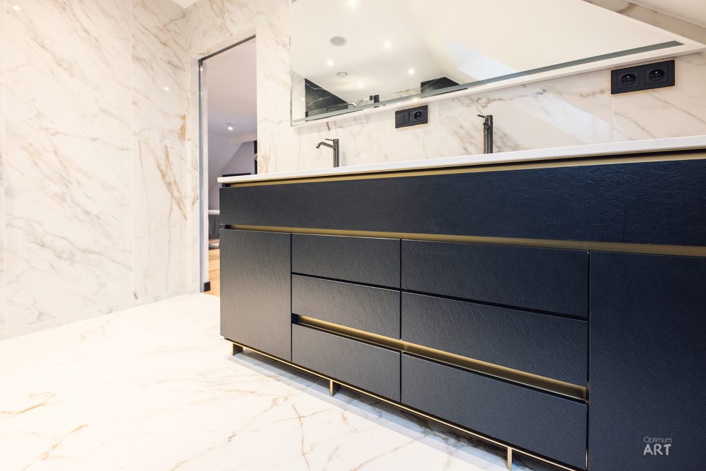 Luksusowa łazienka z granatową szafką podumywalkową z podwójną umywalką i złotymi akcentami. Ściany wykończone jasnym marmurem ze złotymi żyłkami, na suficie zamontowany czarny projektor.