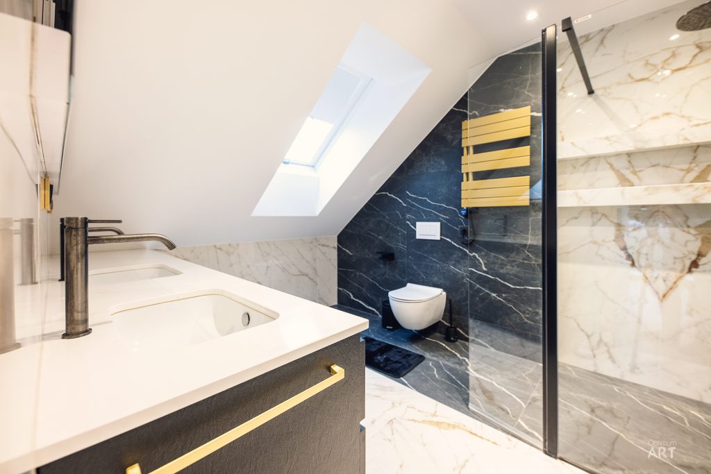 Nowoczesna łazienka na poddaszu z białymi i czarnymi marmurowymi płytkami, umywalką z czarną baterią oraz toaletą w szklanej kabinie. Pomieszczenie ma skośny sufit z oknem dachowym i złote akcenty na meblach.