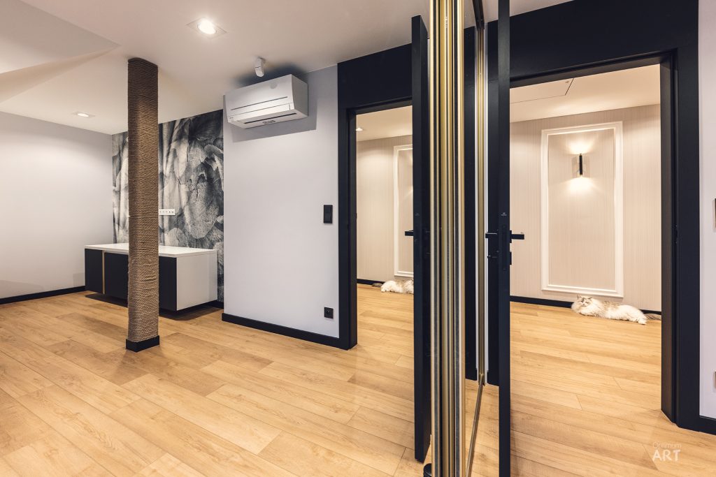 Nowoczesne wnętrze mieszkania z drewnianą podłogą, czarnymi ramami drzwi i okien oraz białymi ścianami. W pomieszczeniu znajduje się kolumna owinięta sznurem, klimatyzator na ścianie oraz widoczne są otwarte drzwi prowadzące do innych pomieszczeń.