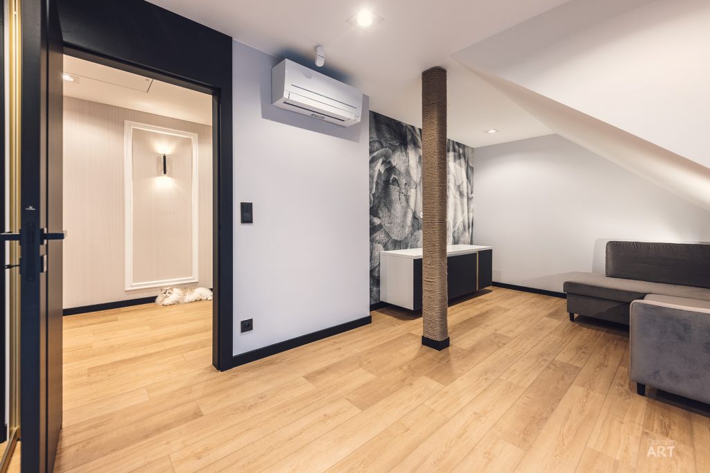 Nowoczesne wnętrze mieszkania z jasnym drewnianym podłożem, białymi ścianami i czarną kolumną owiniętą sznurem. W pomieszczeniu znajdują się szare meble wypoczynkowe, klimatyzator na ścianie oraz przejście do kolejnego pokoju z widocznym białym drzwiami.