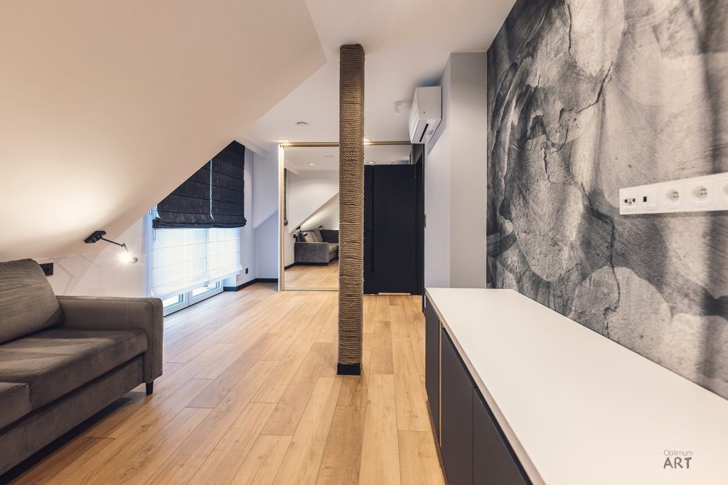 Nowoczesne wnętrze mieszkania na poddaszu z drewnianą podłogą, szarą sofą po lewej stronie i marmurową ścianą po prawej. W centrum znajduje się kolumna owinięta sznurem, a w tle widoczne są ciemne rolety okienne i przejście do kolejnego pomieszczenia.