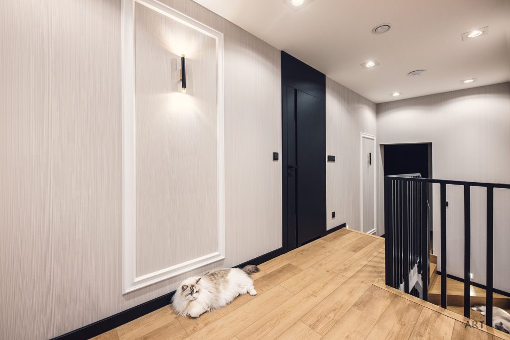 Nowoczesny korytarz z jasnymi ścianami, drewnianą podłogą i czarnymi drzwiami, przy których leży biało-szary kot. Po prawej stronie widoczne są czarne balustrady schodów.