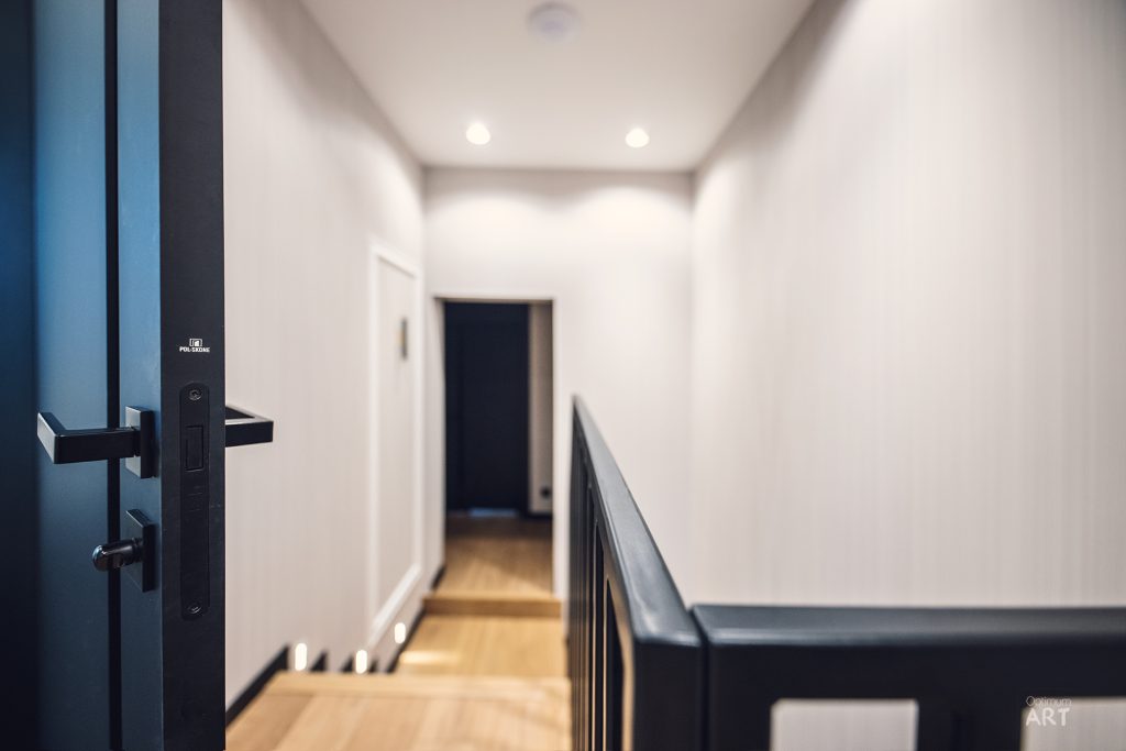 Nowoczesny korytarz w mieszkaniu z białymi ścianami, drewnianą podłogą i czarną balustradą po prawej stronie. Po lewej stronie widoczne są ciemnoszare drzwi z elektronicznym zamkiem.