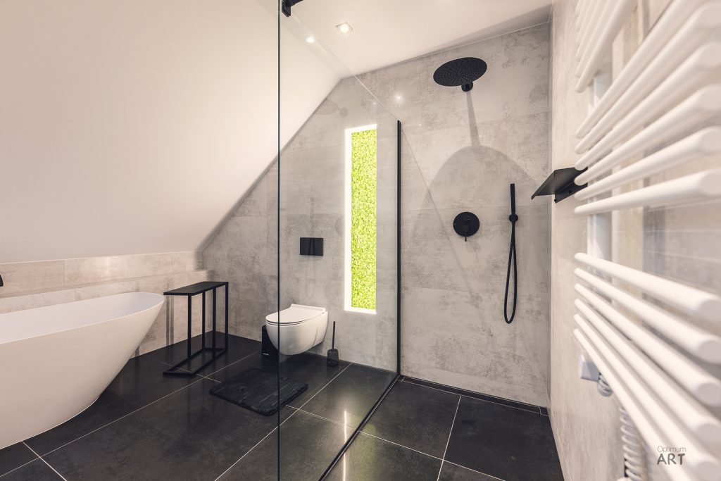 Nowoczesna łazienka na poddaszu z wanną wolnostojącą, prysznicem walk-in ze szklaną ścianką i czarnymi armaturami. Pomieszczenie ma szare ściany betonowe, czarną podłogę oraz biały grzejnik drabinkowy.