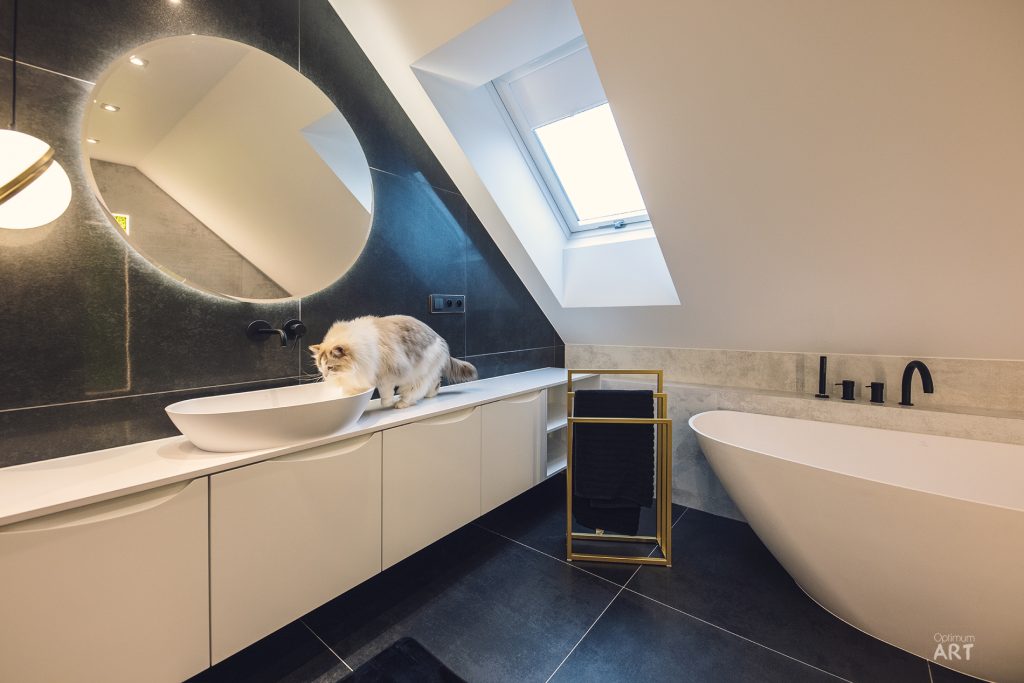 Nowoczesna łazienka na poddaszu z białą wanną wolnostojącą, umywalką nablatową i okrągłym lustrem. Na blacie przy umywalce siedzi puszysty kot o jasnym umaszczeniu.