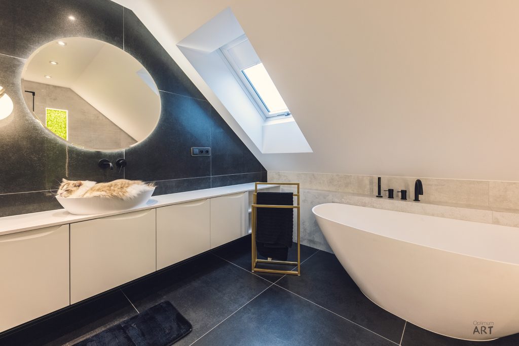Nowoczesna łazienka na poddaszu z białą wolnostojącą wanną, okrągłym lustrem nad umywalką i oknem dachowym. Wnętrze utrzymane w kolorystyce bieli i ciemnego granatu z czarnymi kafelkami na podłodze.
