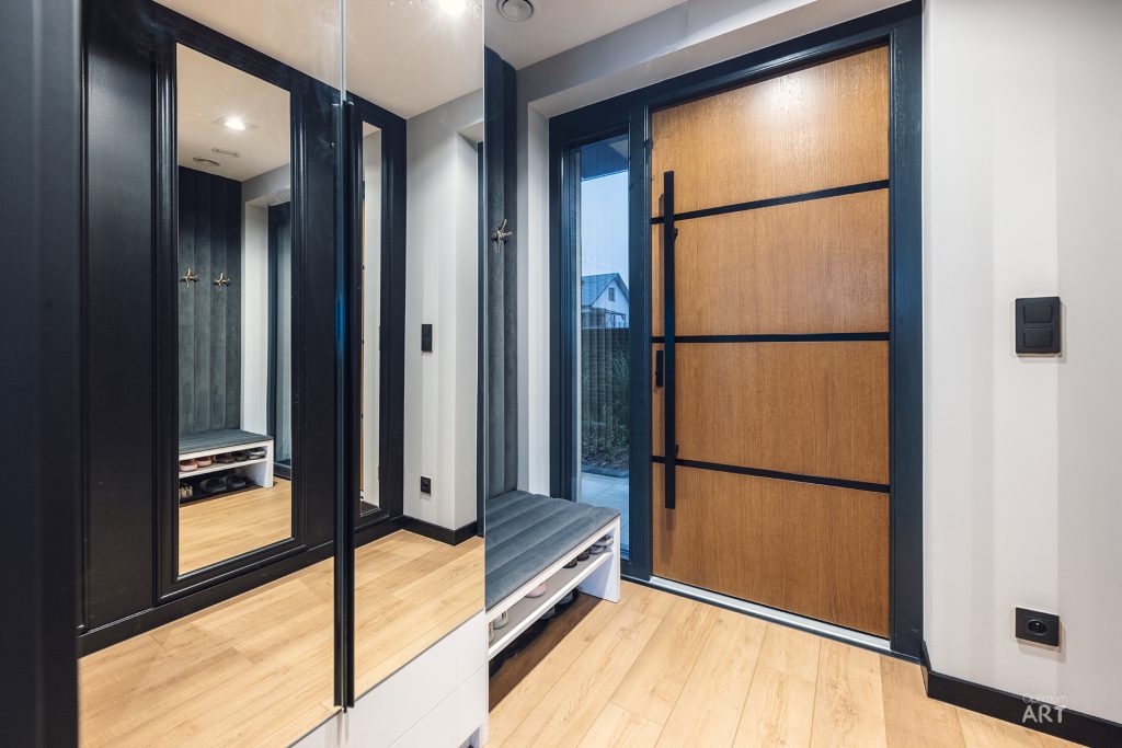 Nowoczesny hol z drewnianą podłogą, dużymi lustrzanymi drzwiami szafy po lewej stronie i drewnianymi drzwiami wejściowymi z czarnymi poziomymi pasami po prawej. W przestrzeni znajduje się także niewielka ławka i widoczne jest odbicie łazienki w lustrze.