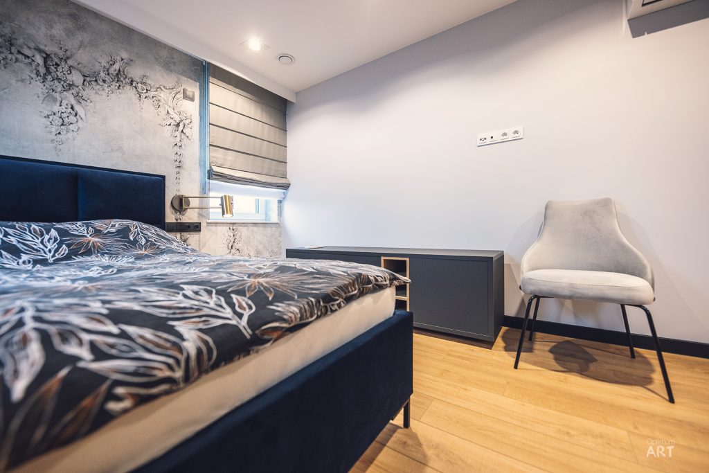 Nowoczesna sypialnia z ciemnoniebieskim łóżkiem i wzorzystą pościelą, szarym fotelem oraz dekoracyjną ścianą z roślinnym motywem. Pomieszczenie ma drewnianą podłogę, białe ściany i okno z roletą.