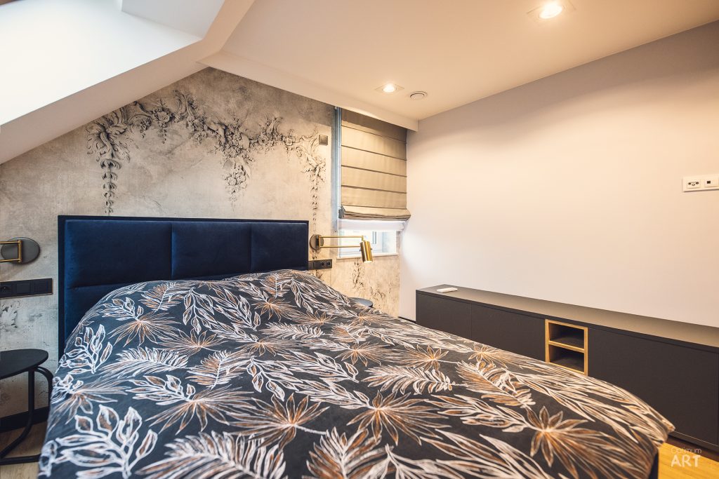 Sypialnia z podwójnym łóżkiem z granatowym zagłówkiem i pościelą w roślinny wzór, umieszczona pod skosem dachu. Na ścianie za łóżkiem widoczna tapeta z motywem roślinnym, a po prawej stronie znajduje się ciemna komoda z półkami.