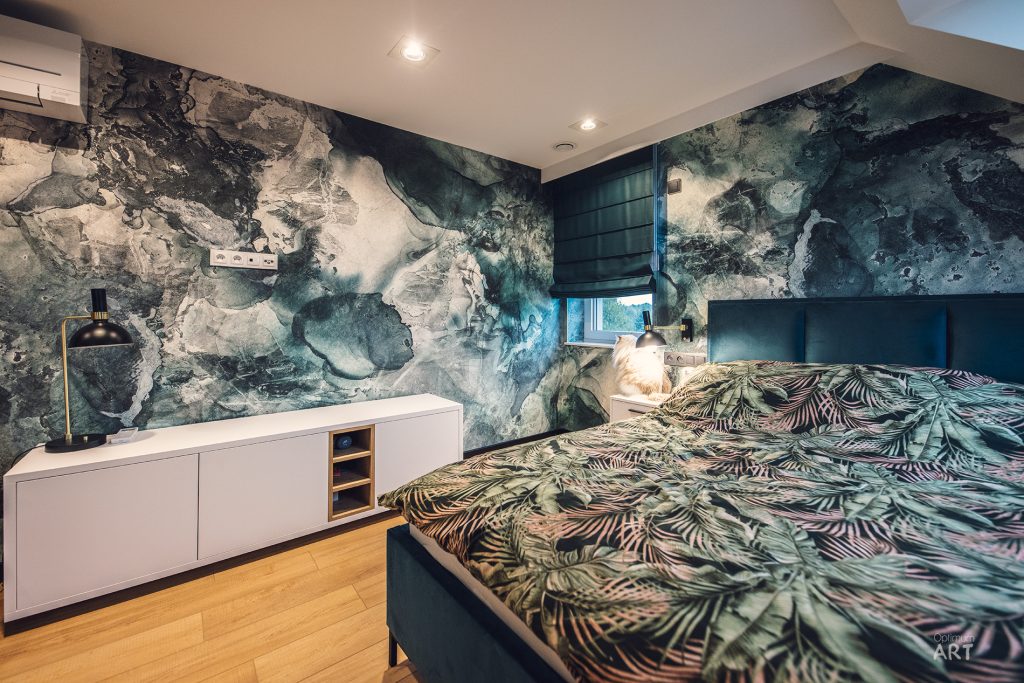 Sypialnia z dużym łóżkiem przykrytym pościelą w tropikalny wzór liści palmowych. Ściany pokryte są artystycznymi tapetami w odcieniach zieleni i szarości przypominającymi abstrakcyjny krajobraz lub marmurowe wzory.