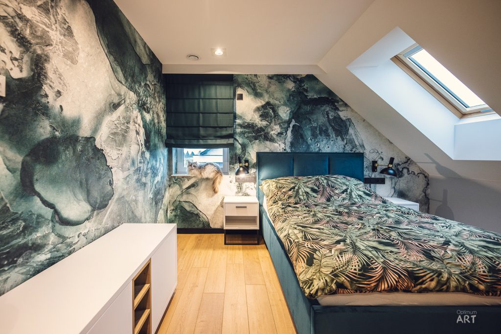 Sypialnia na poddaszu z ukośnym sufitem i świetlikiem dachowym, wyposażona w łóżko z zielonymi pościelami w tropikalne wzory. Ściana za łóżkiem pokryta jest tapetą z motywem oceanicznym w odcieniach zieleni i niebieskiego, a podłoga wykonana z jasnego drewna.
