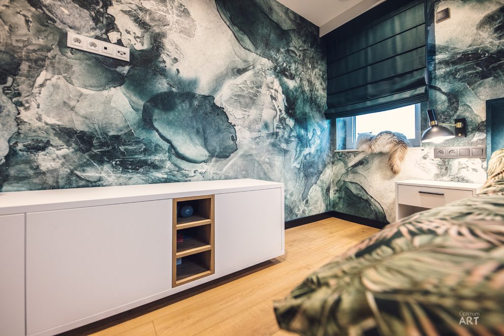 Nowoczesna kuchnia z białymi szafkami dolnymi i spektakularną fototapetą na ścianie przedstawiającą abstrakcyjny wzór w odcieniach niebieskiego i szarego przypominający mapę świata lub formacje skalne. Podłoga wykonana z jasnego drewna, a w prawym dolnym rogu widoczne są liście rośliny doniczkowej.