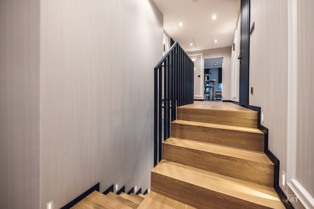 Drewniane schody prowadzą w górę w nowoczesnym wnętrzu z jasną ścianą i czarną metalową balustradą. Na górze schodów widać korytarz z recesyjnym oświetleniem i wejście do innych pomieszczeń.