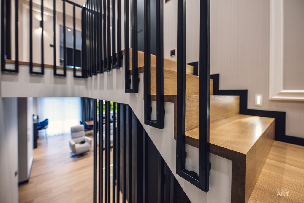 Nowoczesne drewniane schody z czarnymi metalowymi balustradami prowadzą na wyższe piętro. W tle widoczny jest salon z jasnymi meblami i dużymi oknami.