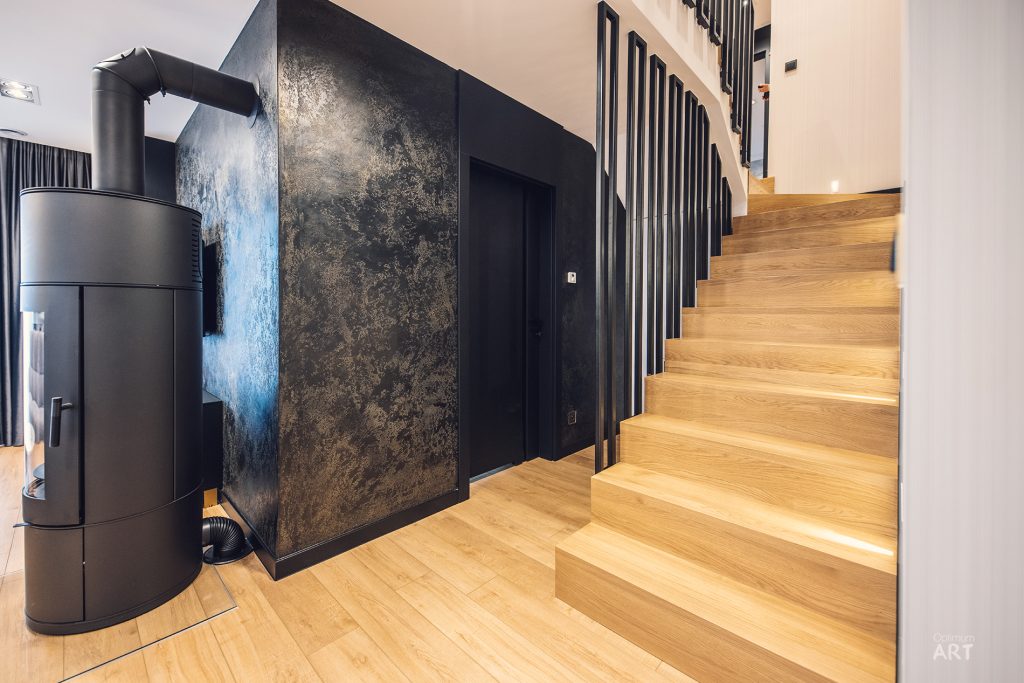 Nowoczesne wnętrze z drewnianymi schodami prowadzącymi na piętro i czarną ścianą z dekoracyjną fakturą. Po lewej stronie znajduje się czarny piec lub kominek z rurą odprowadzającą dym.