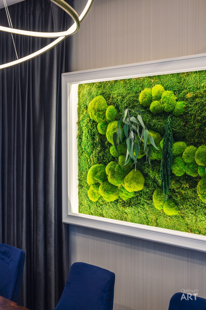 Na ścianie wisi duża rama z żywą zieloną ścianą roślinną z mchem i liśćmi. Poniżej widać fragment niebieskich foteli, a z góry zwisa nowoczesna złota lampa w kształcie pierścieni.