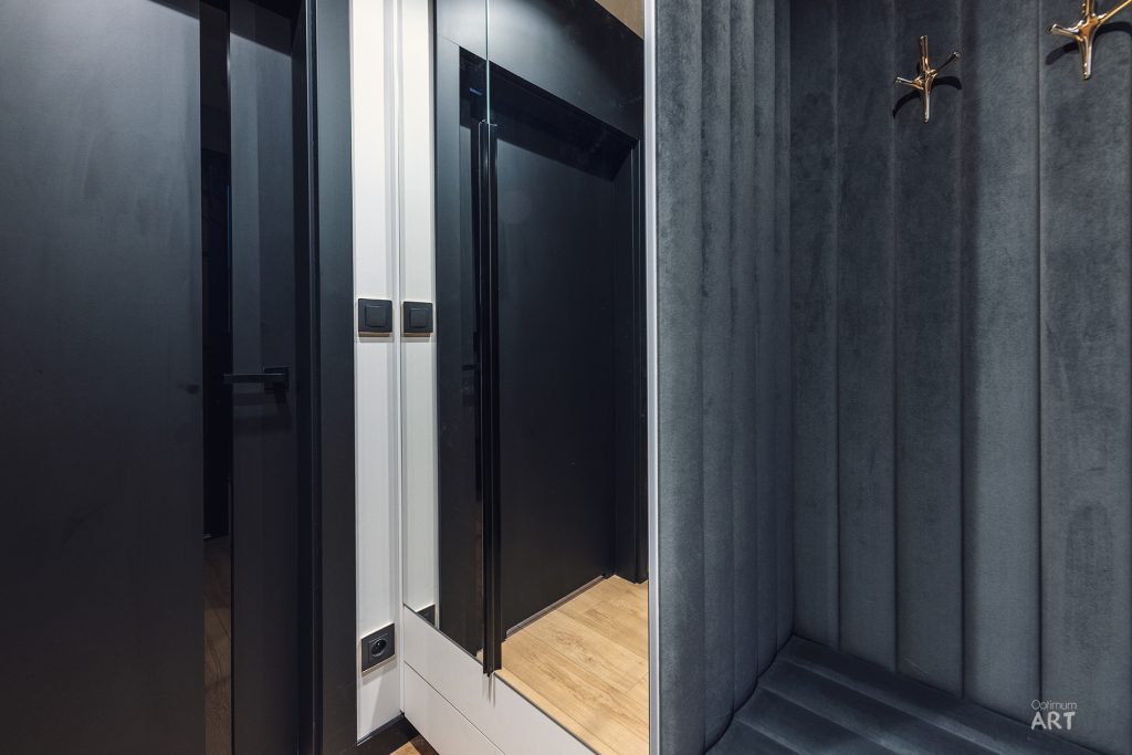 Nowoczesne wnętrze z ciemnoszarymi ścianami i czarnymi drzwiami prowadzącymi do pokoju z jasną drewnianą podłogą. Po prawej stronie znajduje się ściana z panelami w odcieniach szarości z złotymi wieszakami.