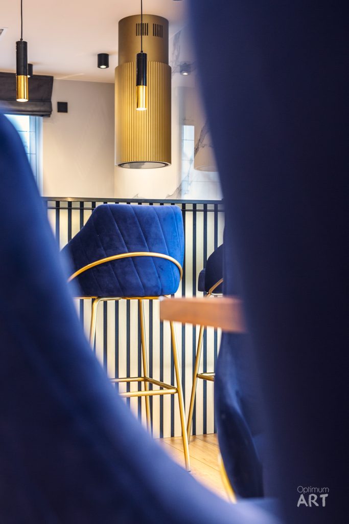 Nowoczesne wnętrze z niebieskim aksamitnym krzesłem barowym przy złotym stole, oświetlone stylową lampą wisząca z drewnianym abażurem. W tle widoczne są czarne metalowe balustrady schodów i jasne ściany z ciepłym oświetleniem.