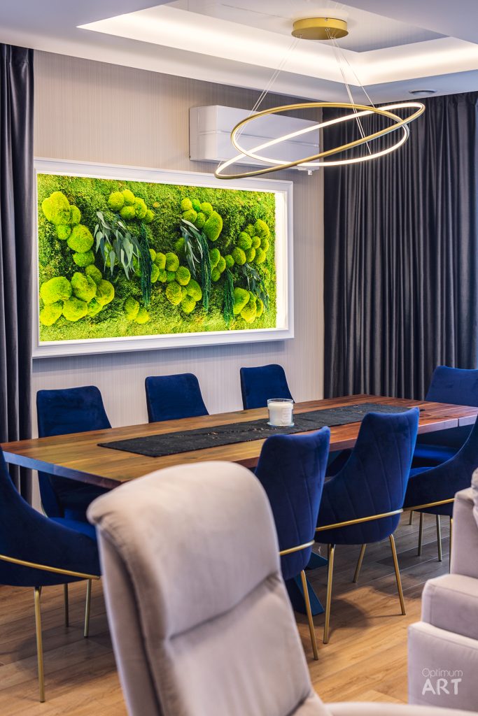 Nowoczesna jadalnia z drewnianym stołem otoczonym niebieskimi krzesłami, nad którym wisi złoty żyrandol w kształcie spirali. Na ścianie umieszczony jest duży obraz z żywą zielenią roślin, a pokój uzupełniają szare zasłony i beżowe fotele.