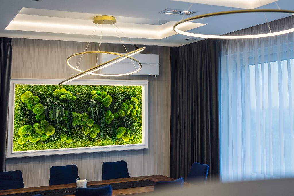 Nowoczesna sala konferencyjna z dużym stołem otoczonym granatowymi krzesłami, na ścianie wisi duży obraz z zieloną roślinnością. Nad stołem znajduje się designerska złota lampa w kształcie przeplatanych kręgów, a po prawej stronie widać okno z zasłonami.