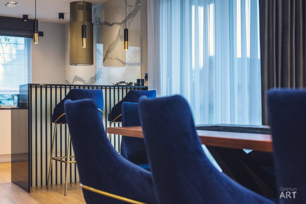 Nowoczesne wnętrze z niebieskimi fotelami przy drewnianym stole oraz wyspą kuchenną z pionowymi czarnymi elementami. W tle widoczna kuchnia z marmurowymi powierzchniami i złotymi lampami wiszącymi, a duże okno z białymi zasłonami wpuszcza naturalne światło.