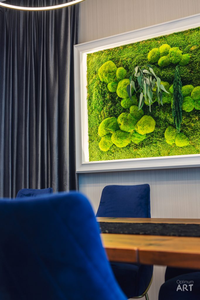 Nowoczesne wnętrze z niebieskimi krzesłami przy drewnianym stole i dużym obrazem z zielonych mchów w białej ramie na ścianie. W tle widoczne są ciemnoszare zasłony i stylowe oświetlenie sufitowe.