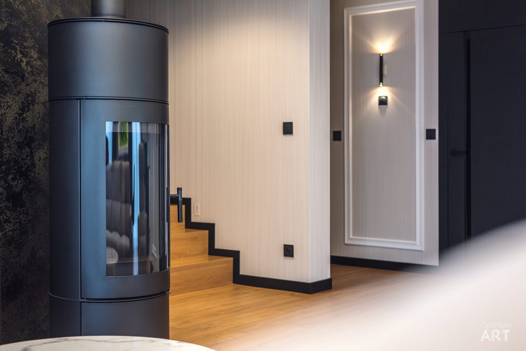 Nowoczesne wnętrze z cylindrycznym kominkiem o szklanej obudowie i ciemnej metalowej ramie, ustawionym na drewnianej podłodze. W tle widoczne są schody oraz elegancko oświetlona wnęka w ścianie z dekoracyjnym panelem.