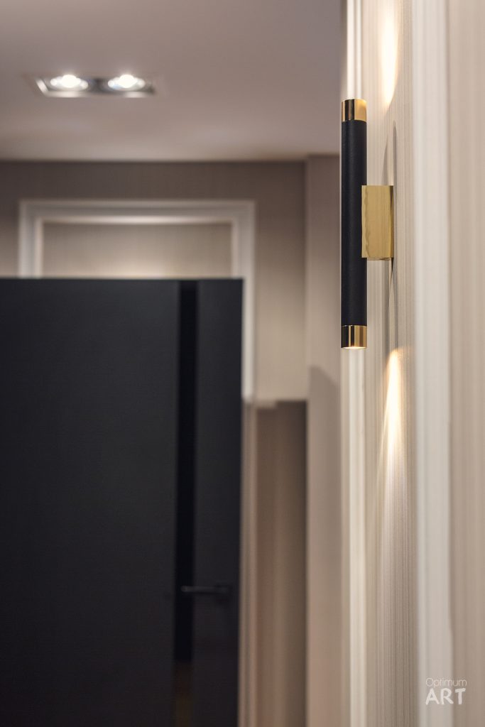 Nowoczesna kinkiet ścienny w kolorze czarnym ze złotymi akcentami, zamontowany na białej ścianie, emituje ciepłe światło w górę i w dół. W tle widoczne jest eleganckie wnętrze z białymi drzwiami i sufitem z wpuszczanymi lampami.