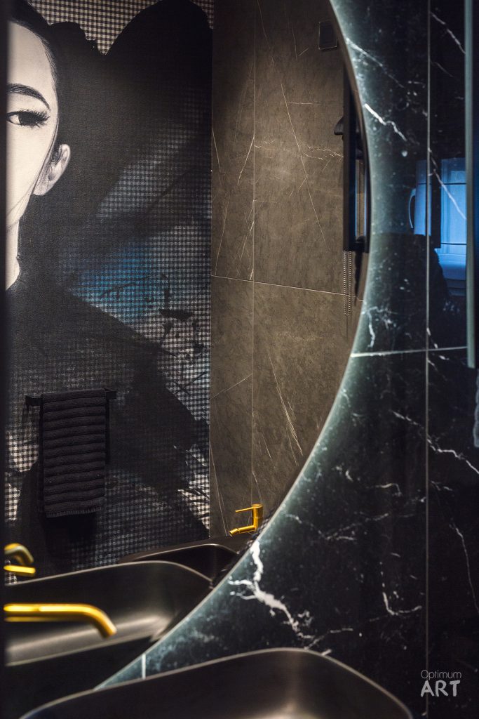 Luksusowa łazienka z czarnym marmurem i mozaiką, w której dominuje duży portret kobiety na ścianie. Wnętrze charakteryzuje się eleganckimi, ciemnymi kolorami z akcentami złota i niebieskiego oświetlenia.