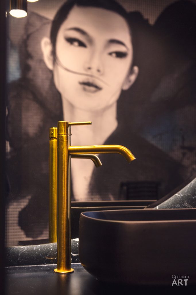 Złota bateria umywalkowa stoi przy ciemnej umywalce na tle dużego portretu młodej kobiety o azjatyckich rysach twarzy namalowanego na ścianie łazienki.